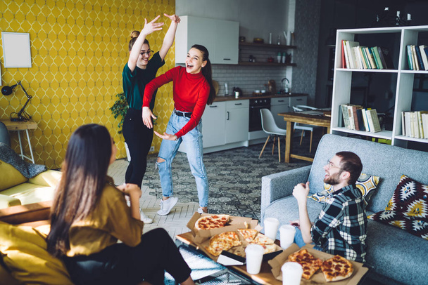 Laughing amused women standing and gesturing while playing charades with friends by table with delivered pizza and disposable cups in sunny room - 写真・画像