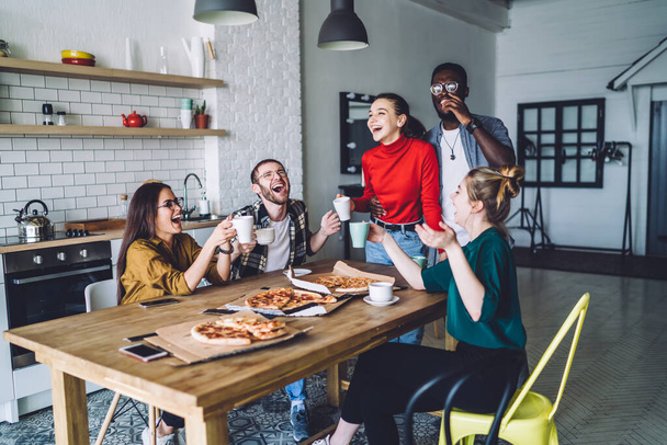Casual multiethnic students with drinks in hands and pizza at table making good cheer and laughing at cozy stylish kitchen  - 写真・画像