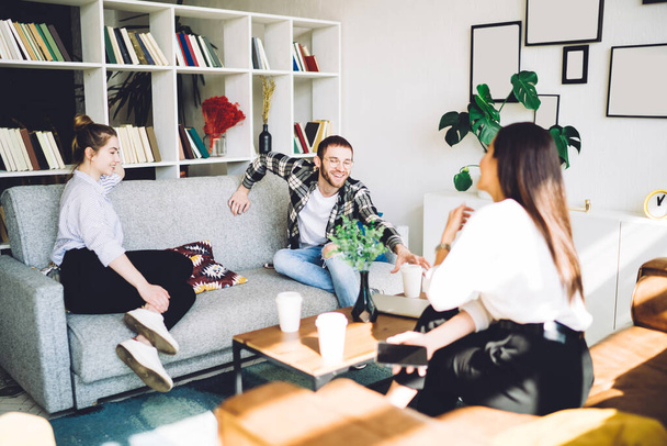 Happy friends sharing news and drinking coffee together while sitting on comfortable sofa at coffee table in modern stylish flat - Photo, Image