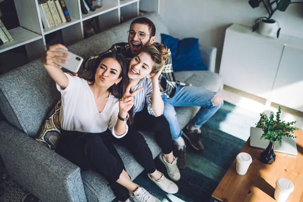 From above dark haired expressive woman taking selfie with happy woman and cheerful man in glasses in cozy modern apartment - Foto, afbeelding