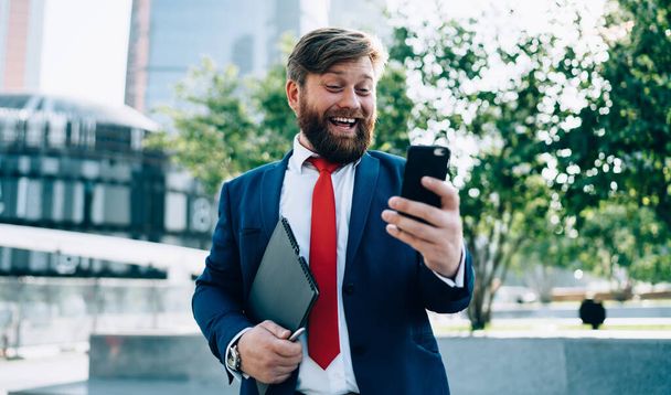 Bright happy man in elegant clothes with red tie having fun talking to coworker chilling in modern park of city - Foto, afbeelding