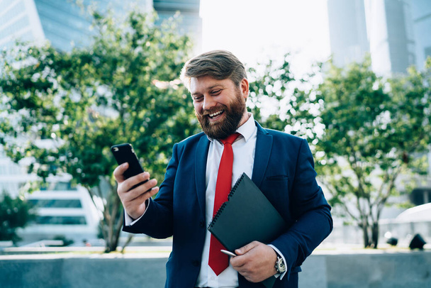 Cheerful bearded male in suit with red tie calling partner via video chat on phone walking in park of modern city - Fotoğraf, Görsel