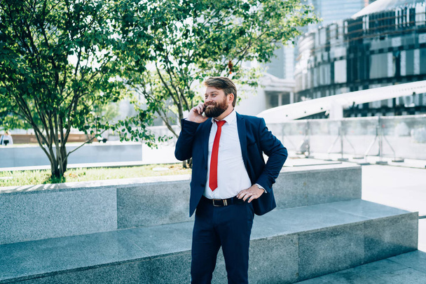 Cheerful bearded man in dark elegant suit and red tie enthusiastically talking on mobile phone in square of business center  - Foto, Imagen