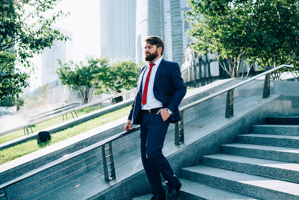 Side view of bearded thoughtful tall businessman in elegant suit and red tie walking with hand in pocket on stairs leaning handle on background of glass building  - Fotografie, Obrázek