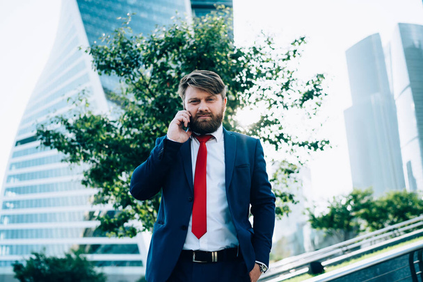 Cheerful elegant man in dark classic suit and red tie listening while speaking on mobile phone and looking at camera with hand in pocket in square of business center  - Фото, изображение