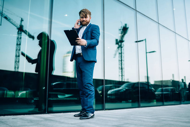 From below joyful adult executive man in elegant blue suit with papers looking at camera and making phone call while standing on street against contemporary building with glass walls - Photo, Image