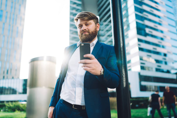 From below of contemporary busy bearded male in formal suit looking at camera with pleasant smile while standing in back lit next to office building leaning on post and using smartphone - Fotó, kép