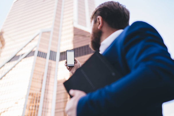 Low angle of blurred formal man with beard in blue suit holding folder in hand and raising hand up with smartphone  - Foto, Bild