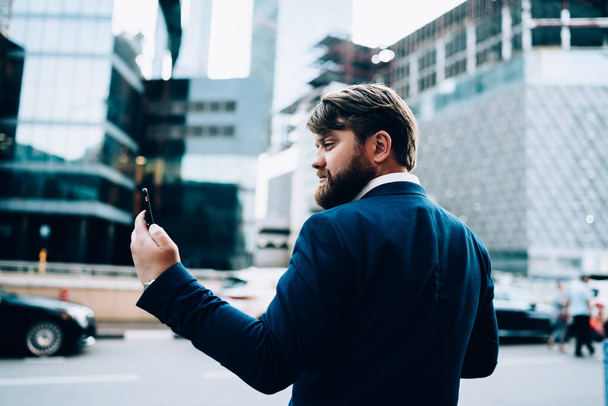 Back view of elegant serious businessman taking selfie on mobile phone looking looking confidently half turn on background of glass business center  - Foto, immagini