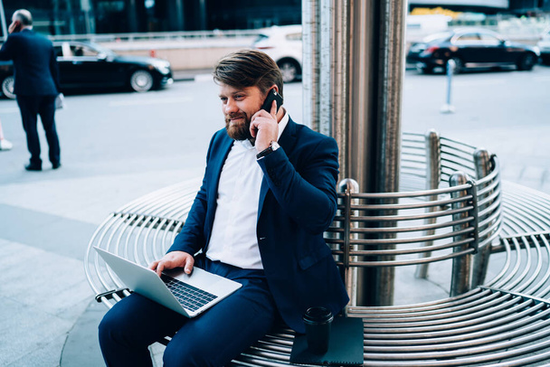 Hombre satisfecho hablando por teléfono y sosteniendo el ordenador portátil mientras está sentado en el banco en medio de la calle ocupada a la luz del día - Foto, imagen
