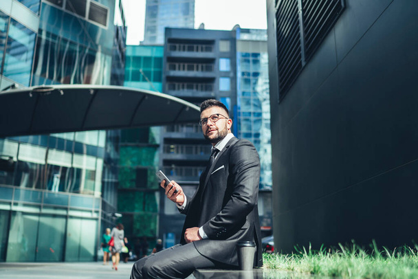 From below confident pleased executive man in glasses and elegant clothes looking away and developing plans for future while sitting on stone bench and interacting with smartphone on street during coffee break against contemporary buildings - Foto, imagen