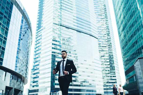 Busy adult businessman in elegant black suit with beverage to go walking along street against blurred contemporary high rise office buildings with glass facades in downtown - Fotó, kép