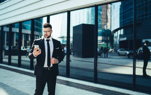 Male executive in trendy eyeglasses and business style clothing surfing on mobile phone while standing among modern buildings at downtown - Фото, зображення