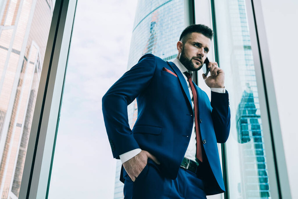 From below confident well dressed man having business discussion on phone while standing with hand in pocket against window wall in contemporary office - Photo, image