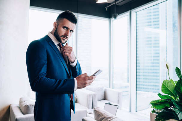 Compañero de trabajo masculino preocupado en traje formal azul frotando la barbilla en pensamientos mientras navega en el teléfono móvil y mirando la cámara en la oficina   - Foto, imagen