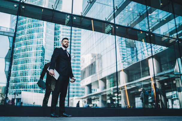 Low angle of serious entrepreneur with tablet and smartphone looking away and thinking while standing on pavement outside modern building with glass walls - 写真・画像