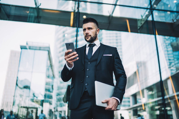 Low angle of serious guy in suit carrying tablet and looking away while standing on street of modern city and browsing smartphone - Фото, зображення
