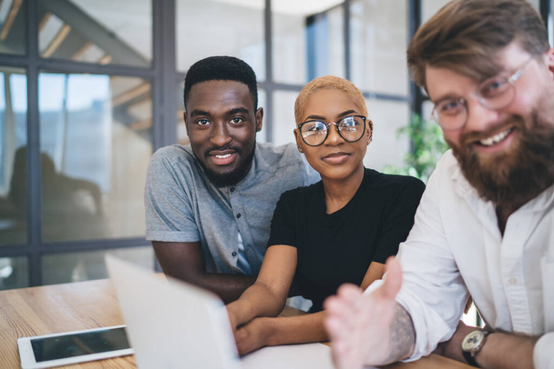 Pleased group of multiracial male and female friends in causal outfit and with glasses sitting at desk surfing laptop looking at camera  - Foto, immagini