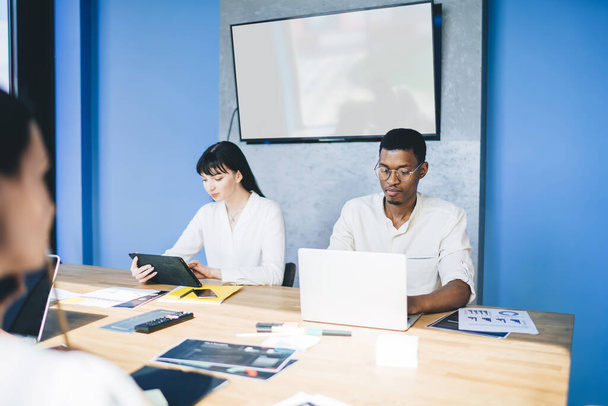 Серйозні багатоетнічні колеги переглядають ноутбук і планшет, сидячи за конференц-столом з графіками і паперами в сучасному офісі разом
 - Фото, зображення