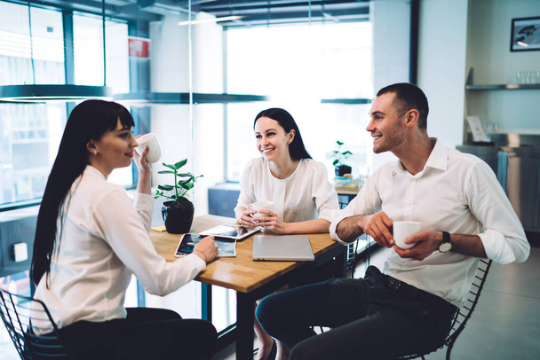 Cheerful office employees having conversation and looking at each other while drinking morning cup of coffee and sitting in modern cafe on wooden table with gadgets - Photo, image
