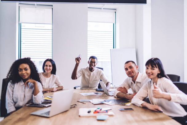 Successful ethnic employee sitting in bright office and playing with paper airplane while cheerful and merry coworkers looking at camera - Foto, Imagem