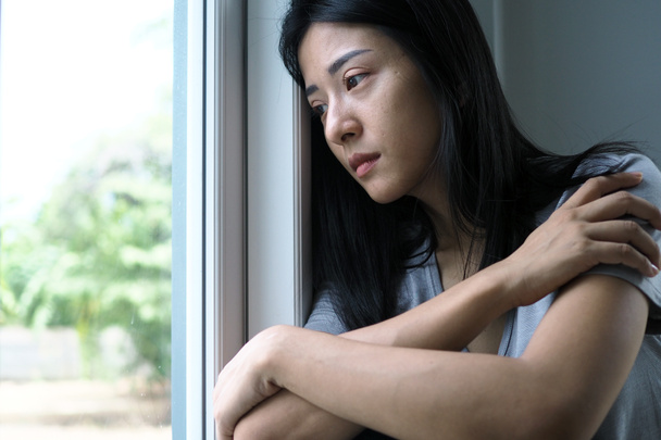 Ασιάτισσα που κάθεται μέσα στο σπίτι και κοιτάει έξω από το παράθυρο. Γυναίκα μπερδεμένη, απογοητευμένη, λυπημένη και αναστατωμένη. - Φωτογραφία, εικόνα