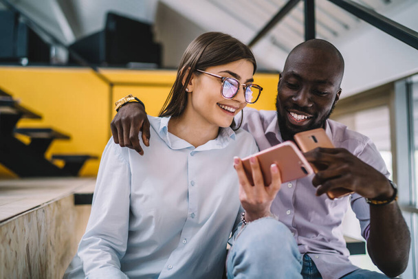 Young African American male embracing cheerful girlfriend and commenting funny photo in internet while sitting together and using mobile phones - Fotografie, Obrázek