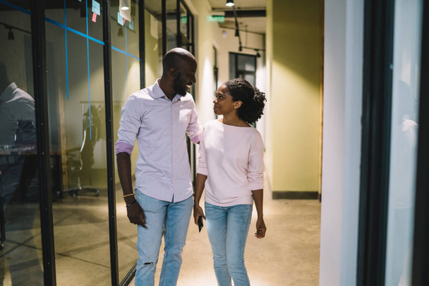 Young content black female with phone in casual clothes resting from work chatting with laughing African american colleague hugging in modern office - Photo, image