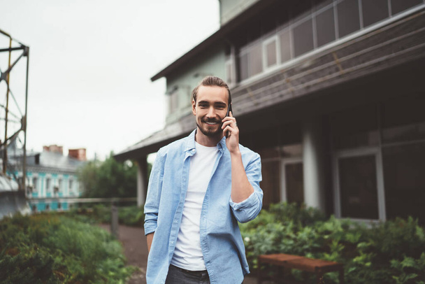 Cheerful young guy in casual outfit looking at camera and answering phone call while standing in garden of suburban house - Fotografie, Obrázek