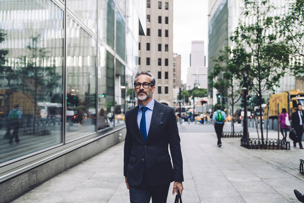 Bem sucedido confiante preparado cinza-barbudo homem de negócios de meia idade em terno preto caro e óculos andando confiantemente na rua de Nova York
 - Foto, Imagem