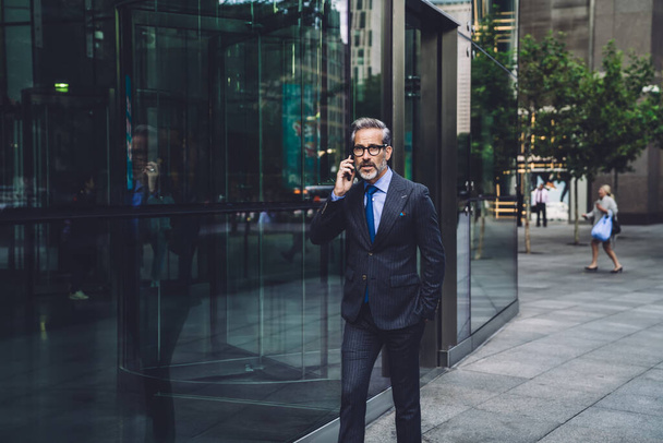 Handsome fashionable businessman with beard in stylish dark business suit walking talking on phone near office building in city street in New York - Foto, Imagem