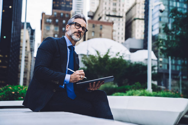 Side view of middle aged businessman in glasses with legs crossed holding tablet with stylus while sitting on bench and looking away against New York buildings - 写真・画像