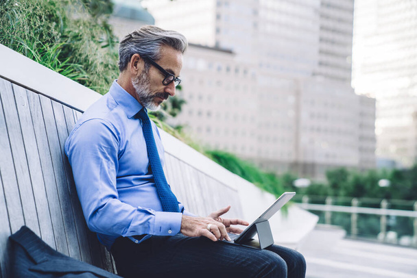 Side view of mature male in glasses typing on tablet with keyboard while sitting on wooden bench with business suit against background of New York buildings - Фото, изображение