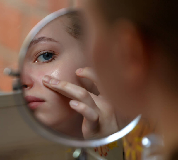 Girl teenager checking pimples on her face in front of round mirror - Фото, изображение