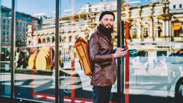 Young bearded male wearing trendy brown leather jacket yellow backpack and knitted black hat browsing smartphone while standing near glass wall of building  and looking away - Фото, зображення
