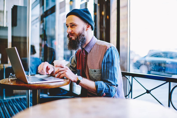 Young content bearded man dressed in stylish casual shirt and knitted hat working with laptop and drinking coffee in cafe at daytime - Foto, imagen