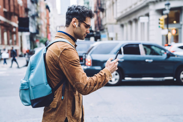 Side view of serious Hispanic young man in eyeglasses wearing brown suede jacket with backpack and earphones texting on cellphone and listening music while wandering around New York city - 写真・画像