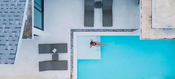Aerial top view, carefree female tourist in red swimsuit relaxing in luxury modern villa swimming pool during summer vacation holidays. Fashionable woman resting in rental property - Zdjęcie, obraz