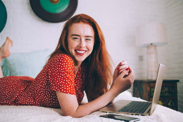 Smiling optimistic red-haired woman in polka dot dress lying on soft bed while using smartphone and laptop in minimalistic room and looking at camera - Photo, Image