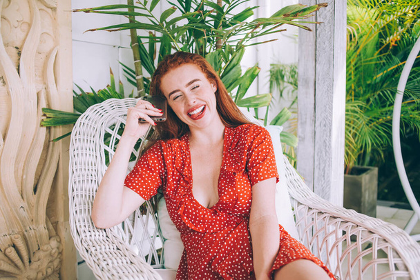 Laughing attractive adult red haired female in casual red dotted dress and red lipstick relaxing in chair with smartphone on apartment decking and looking at camera - Fotografie, Obrázek