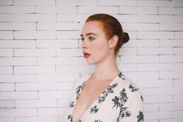 Side view of young beautiful pensive ginger woman in pretty dress standing in front of white brick wall looking away - Foto, Imagem