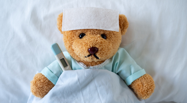 Плюшевый мишка болен на кровати с высокой температурой. На лбу лихорадка, снижающая температуру. - Фото, изображение