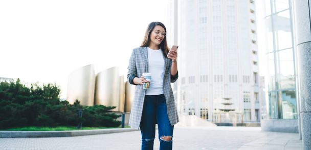 From below trendy young pleased female in casual clothes with coffee to go concentrating on screen and interacting with smartphone while walking along street against blurred modern high rise buildings - 写真・画像