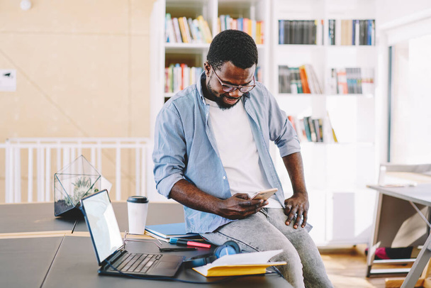 Active African American man in glasses working comfortably placed on table with laptop and documents and surfing mobile phone in light cabinet  - Foto, Imagen