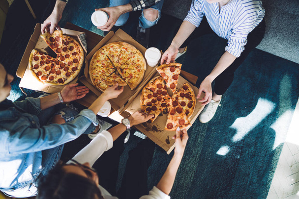 Top view of anonymous people with takeaway beverage taking delicious pizza from boxes while sitting around table during friends gathering in cozy home - Φωτογραφία, εικόνα