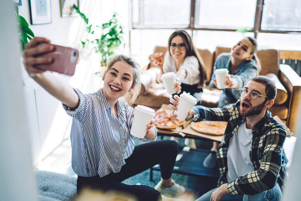 Smiling young woman with beverage to go using smartphone to take selfie with happy friends during party in cozy living room - Photo, image
