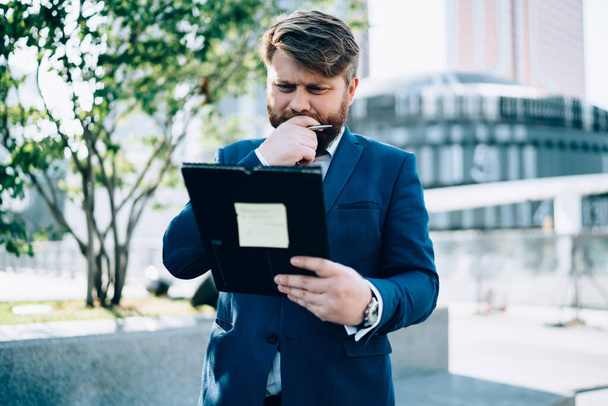 Bearded thoughtful businessman in suit standing on street in business center holding folder reading paper and rubbing chin in dilemma  - Фото, изображение