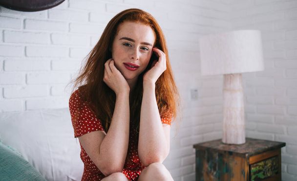 Happy and glad red haired woman in red polka dot dress sitting on bed in bright room with minimalistic interior and looking at camera - Zdjęcie, obraz