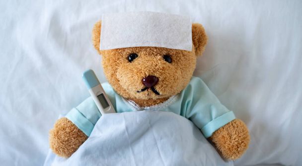 El oso de peluche está enfermo en la cama con fiebre alta. Hay una sábana reductora de fiebre en la frente. - Foto, Imagen
