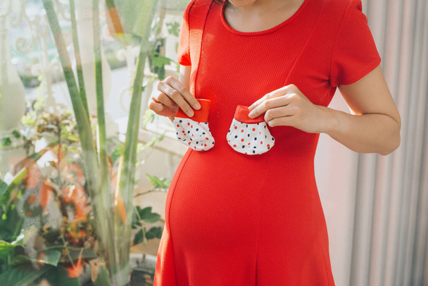 Mujer embarazada sosteniendo pequeños calcetines de bebé relajándose en casa en la sala de estar
. - Foto, Imagen